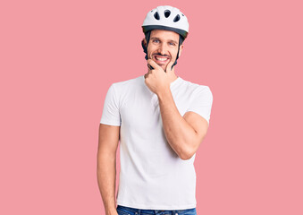 Young handsome man wearing bike helmet looking confident at the camera smiling with crossed arms and hand raised on chin. thinking positive.