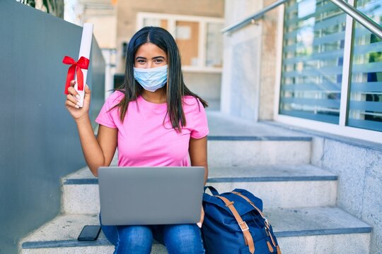 Young latin student girl wearing medical mask using laptop and holding diploma at university.