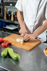 Hands of young male chef cutting fresh onions on wooden board by table
