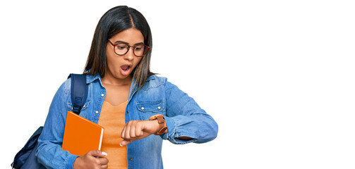 Young latin girl wearing student backpack and holding books looking at the watch time worried,...