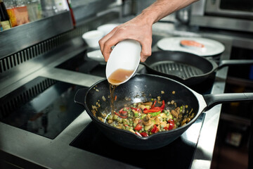 Hand of young chef pouring apple juice into frying pan with chopped ingredients