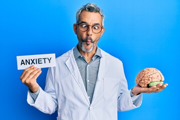 Middle age grey-haired man wearing doctor coat holding brain and anxiety message making fish face...