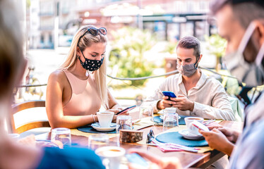 Young friends using mobile smart phones at coffee bar - New normal lifestyle concept with people wearing face mask and checking contact tracing app - Bright filter with focus on left woman