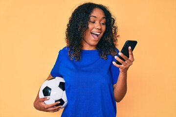 Young african american woman holding football ball looking at smartphone celebrating crazy and amazed for success with open eyes screaming excited.