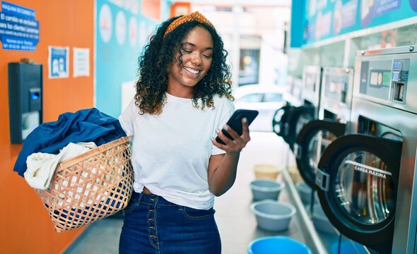 Young african american woman with curly hair smiling happy doing chores at the laundry using phone