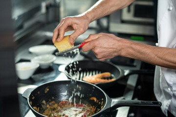 Hands of young chef grating cheese into frying pan with chopped vegetables