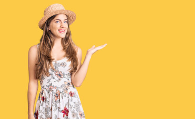 Beautiful caucasian young woman wearing summer hat smiling cheerful presenting and pointing with palm of hand looking at the camera.