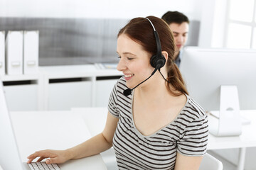 Naklejka premium Casual dressed young woman using headset and computer while talking with customers online. Group of operators at work. Call center, business concept