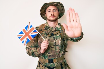 Young caucasian man wearing camouflage army uniform holding united kingdom flag with open hand doing stop sign with serious and confident expression, defense gesture