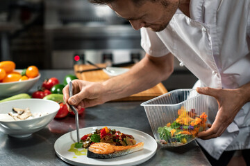 Young male chef decorating roasted vegetables on piece of fried salmon on plate - obrazy, fototapety, plakaty