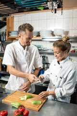 Happy professional chef passing sharp knife to trainee slicing fresh zucchini