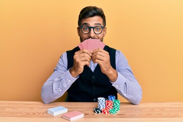 Handsome hispanic croupier man sitting on the table with poker chips and cards smiling and laughing...