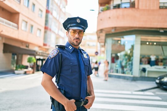 Young handsome hispanic policeman wearing police uniform. Standing with serious expression at town street.