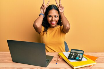 Young african american girl working at the office with laptop and calculator posing funny and crazy with fingers on head as bunny ears, smiling cheerful - obrazy, fototapety, plakaty