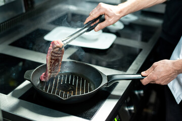 Hands of young chef holding half-roasted piece of meat over hot grill frying pan