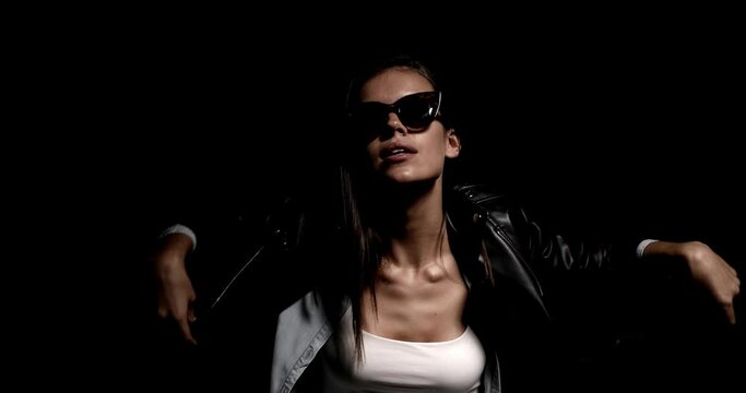 cool young woman wearing leather jacket and sunglasses moving and dancing, taking down glasses, arranging jacket and promoting street style on black background