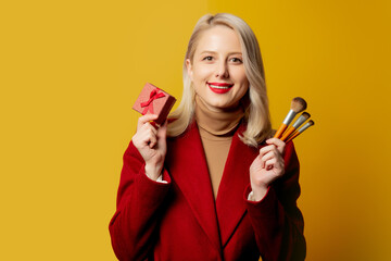 Beautiful woman in red coat with brushes and gift box on yellow background