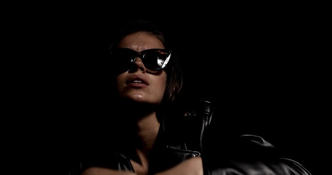 young mysterious woman wearing leather jacket and sunglasses, posing in a fashion light, holding arms in a cool manner, arranging hair and grimacing on black background