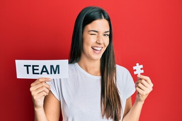 Young brunette woman holding team paper and piece of puzzle as teamwork winking looking at the camera with sexy expression, cheerful and happy face. - obrazy, fototapety, plakaty