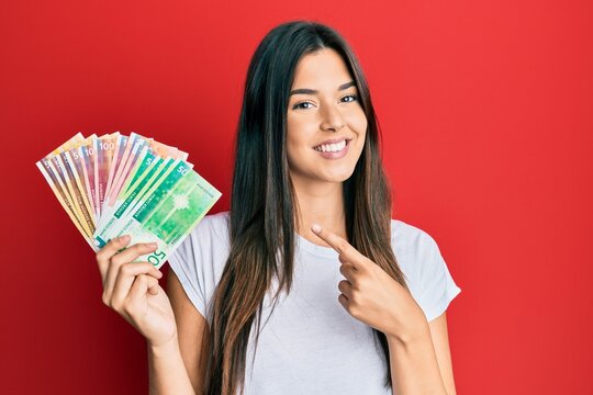 Young brunette woman holding norwegian krone banknotes smiling happy pointing with hand and finger