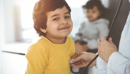 Woman-doctor examining a child patient by stethoscope in sunny clinik. Cute arab boy at physician appointment