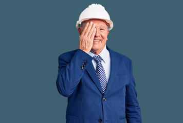 Senior handsome grey-haired man wearing suit and architect hardhat covering one eye with hand, confident smile on face and surprise emotion.