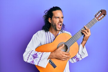 Fototapeta premium Young handsome man with long hair playing classical guitar angry and mad screaming frustrated and furious, shouting with anger. rage and aggressive concept.