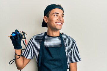Young handsome african american man tattoo artist wearing professional uniform and gloves holding tattooer machine smiling looking to the side and staring away thinking.