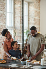 Happy young African man cutting bread for sandwiches by table in the kitchen