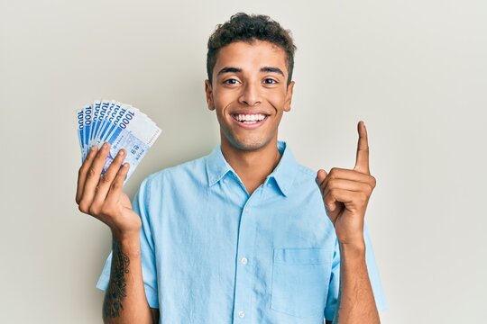 Young handsome african american man holding 1000 south korean won banknotes smiling with an idea or question pointing finger with happy face, number one