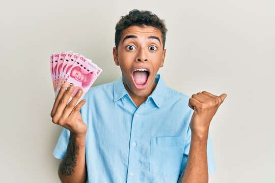 Young handsome african american man holding 100 yuan chinese banknotes pointing thumb up to the side smiling happy with open mouth