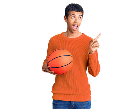 Young african amercian man holding basketball ball pointing thumb up to the side smiling happy with open mouth