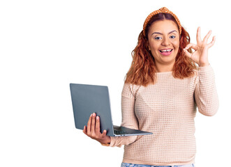 Young latin woman holding laptop doing ok sign with fingers, smiling friendly gesturing excellent symbol