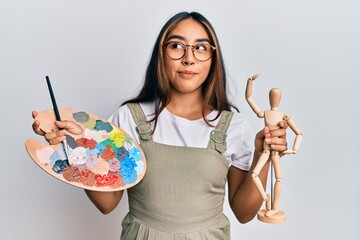 Young latin woman artist holding palette and manikin smiling looking to the side and staring away thinking.