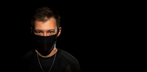 The guy in black clothes and a black medical mask on a black background looks at the camera. Close-up. copy space