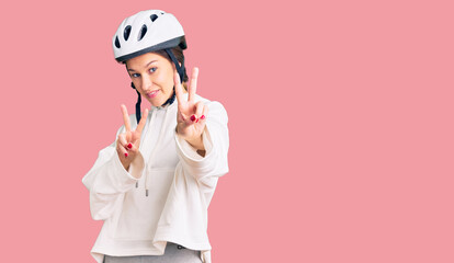 Beautiful brunette young woman wearing bike helmet and sporty clothes smiling looking to the camera showing fingers doing victory sign. number two.