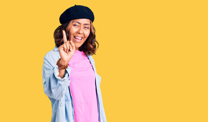 Young beautiful mixed race woman wearing french look with beret showing and pointing up with finger number one while smiling confident and happy.