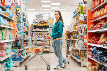 A young pretty Caucasian woman in casual clothing poses with a grocery cart in the aisle of a store. The concept of shopping and buying food