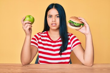 Young asian girl comparing burger and healthy apple clueless and confused expression. doubt concept.