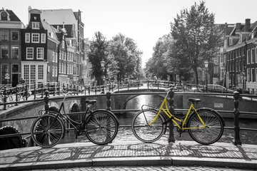 Foto op Plexiglas Fiets A picture of a lonely yellow bike on the bridge over the channel in Amsterdam. The background is black and white.