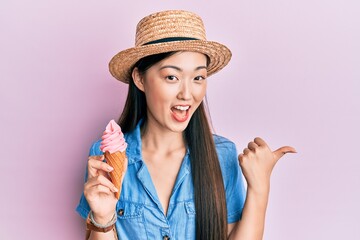 Young chinese woman wearing summer hat holding ice cream pointing thumb up to the side smiling...