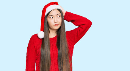 Young chinese woman wearing christmas hat confuse and wondering about question. uncertain with doubt, thinking with hand on head. pensive concept.