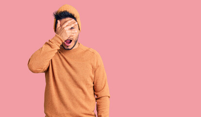 Handsome latin american young man wearing winter sweater and wool hat peeking in shock covering face and eyes with hand, looking through fingers with embarrassed expression.