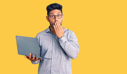 Handsome latin american young man working using computer laptop covering mouth with hand, shocked and afraid for mistake. surprised expression