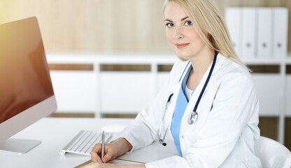 Woman-doctor at work while sitting at the desk in sunny clinic. Blonde cheerful physician filling up medication history record form