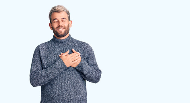 Young handsome blond man wearing casual sweater smiling with hands on chest with closed eyes and grateful gesture on face. health concept.