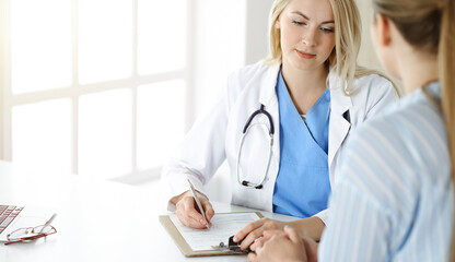 Woman-doctor and patient sitting and talking in sunny clinic. Blonde therapist is cheerfully smiling