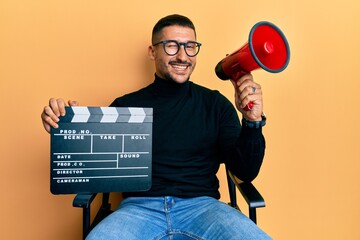 Handsome man with tattoos holding video film clapboard and megaphone winking looking at the camera...