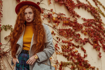 Outdoor autumn fashion portrait of elegant redhead woman wearing orange hat, yellow turtleneck, grey coat, blue checkered trousers, golden wrist watch, holding handbag. Copy, empty space for text