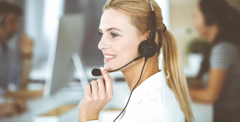 Foto op Plexiglas Blonde business woman using headset for communication and consulting people at customer service office. Call center. Group of operators at work © Iryna